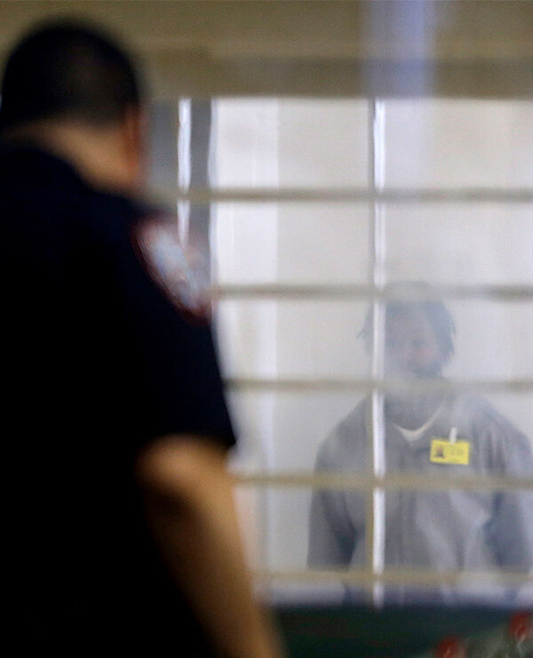 An incarcerated prison looking at a corrections officer separated by bars and a thick plastic barrier.