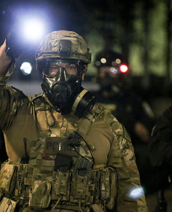 A group of militarized federal agents with an agent in the center shining a bright flashlight toward the viewer.