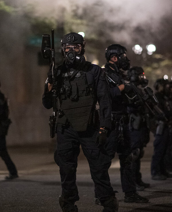 Police in Portland wear riot gear and armed with guns as smoke fills the streets.