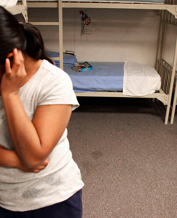 A migrant detainee stands facing away from bunks.