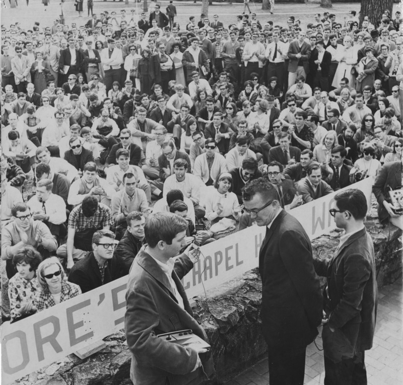 Black and white photo of three men standing in front of a crowd of students.