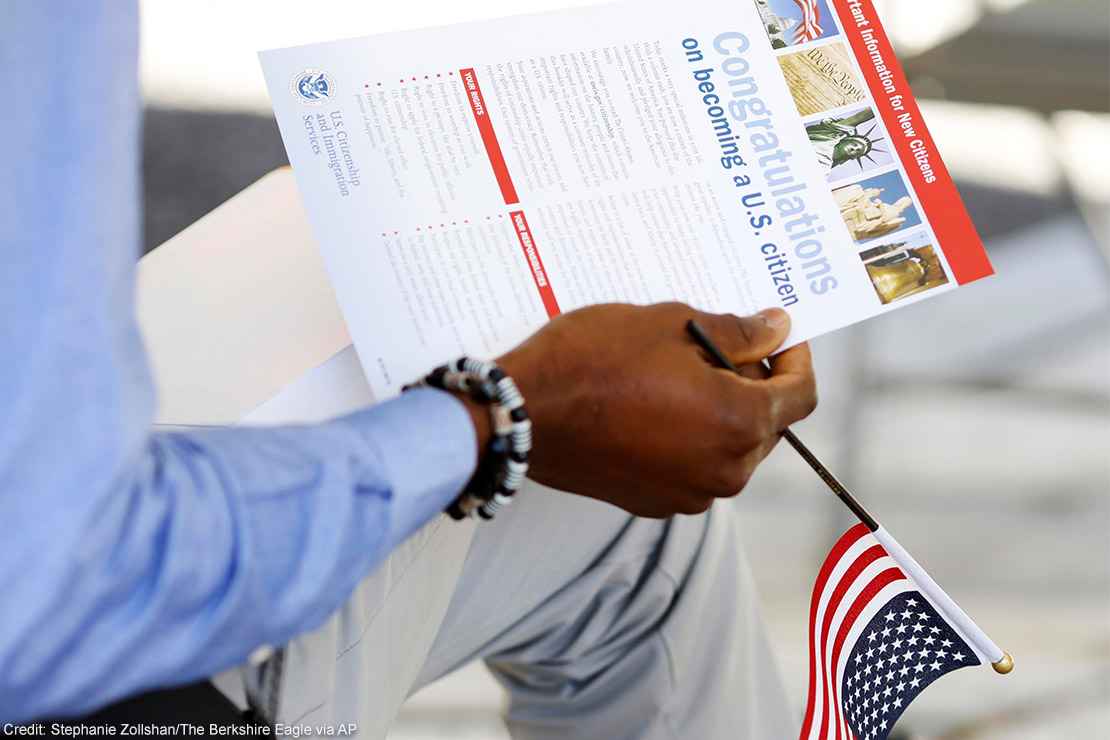 The hand of a seated person holding a miniature U.S. flag and immigration information at a naturalization ceremony.