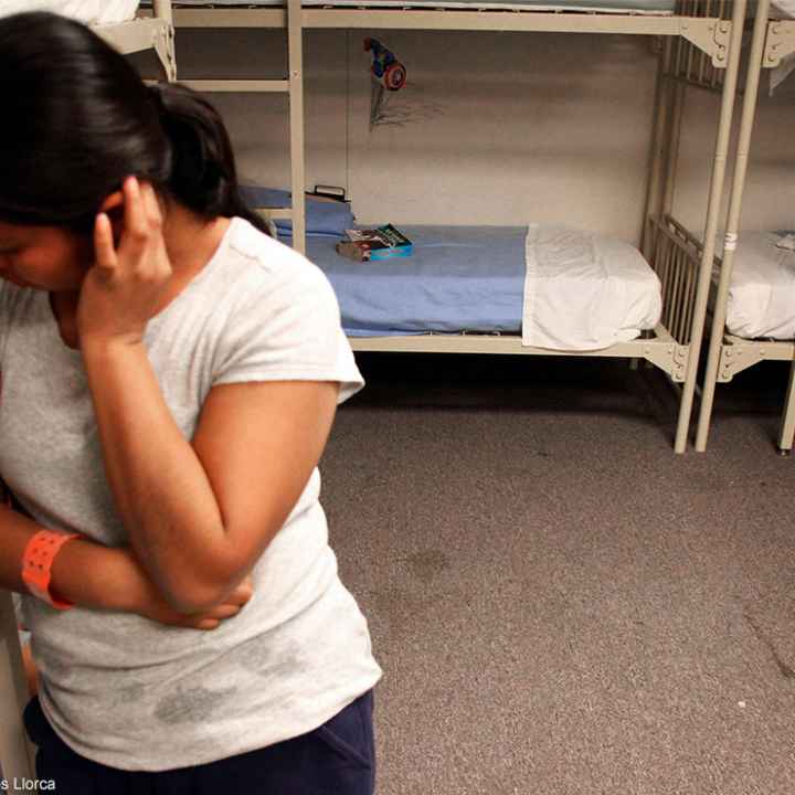 A migrant detainee stands facing away from bunks.